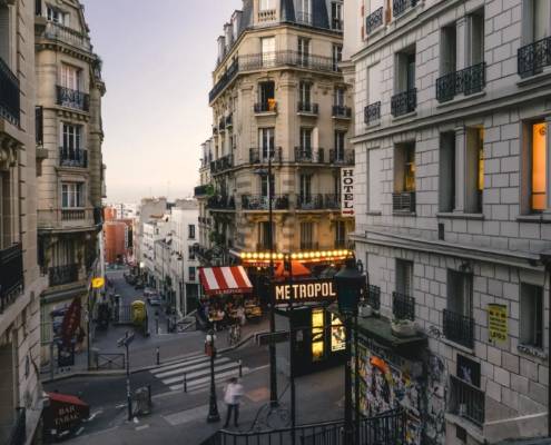 Vue panoramique d'un quartier pittoresque de Paris avec des bâtiments historiques aux façades colorées, des rues pavées animées de passants et de petits cafés, et la Tour Eiffel en arrière-plan sous un ciel bleu clair.