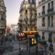 Vue panoramique d'un quartier pittoresque de Paris avec des bâtiments historiques aux façades colorées, des rues pavées animées de passants et de petits cafés, et la Tour Eiffel en arrière-plan sous un ciel bleu clair.