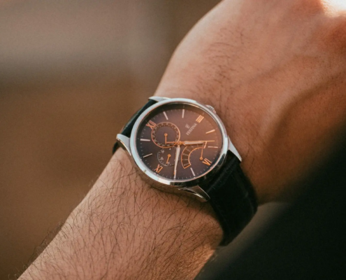 Photo en gros plan d'une montre élégante au cadran argenté et au bracelet en cuir noir, portée sur le poignet gauche d'une personne, avec un fond flou mettant en évidence les détails de la montre